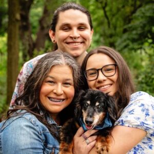 D. Ritchie, Director of Operations, stands smiling at the top center of the image with long dark hair pulled back, wearing a floral shirt and facial piercings. They are flanked by their mother (left), who has long gray-streaked hair and is smiling with braces, and their sister (right), who wears glasses and a white floral blouse. The family’s small black and brown dog is at the bottom center, held lovingly in their arms. The photo is taken outdoors with a green, wooded background.