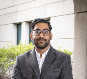 A young man with short, dark hair, a beard, and round glasses stands outdoors in front of a light-colored building wall and green shrubs. He is wearing a dark gray blazer over a white button-down shirt and is smiling slightly with his arms loosely at his sides. The overall...				</div>
			</div>
		</div>
		<div class=